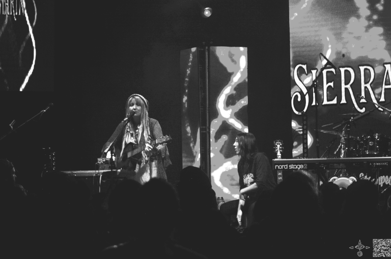 
Sierra Marin performing onstage with a guitar and a keyboard loop station. Green Mystic and Enchanting Logo of her name displayed on display. Black and White photo.
