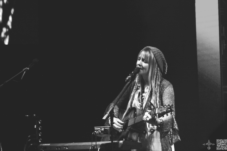 Sierra Marin performing onstage with a guitar and a keyboard loop station. Green Mystic and Enchanting Logo of her name displayed on display. Black and White photo.