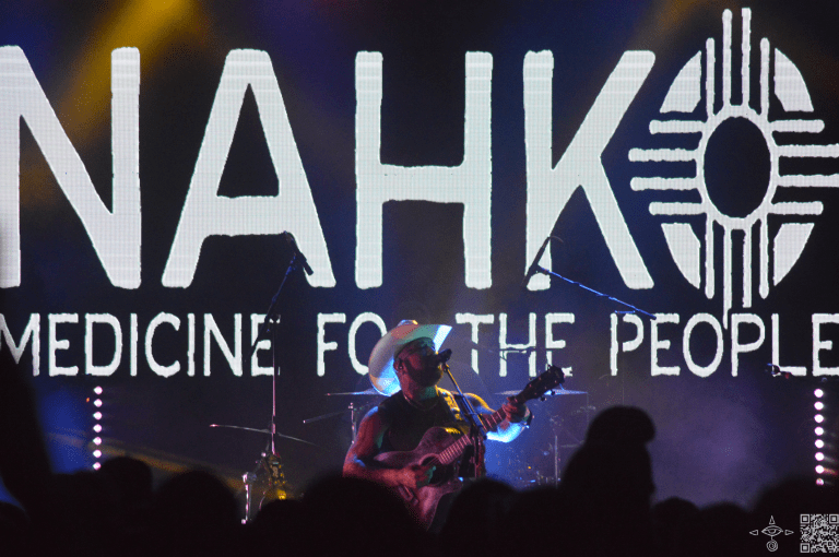 Nahko Performing on stage with a Western Hat
Nahko Looking left with a Guitar, Nahko Logo in white letters, A scanable code logo in the bottom right corner