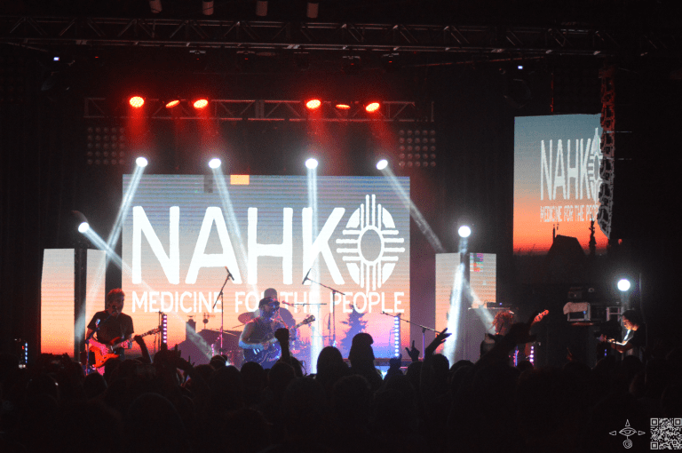 Zoomed out perspective of the stage, white beaming lights, colors of a sunset on screen, Nahko Logo, in white letters, band and crowd, A scanable code logo in the bottom right corner