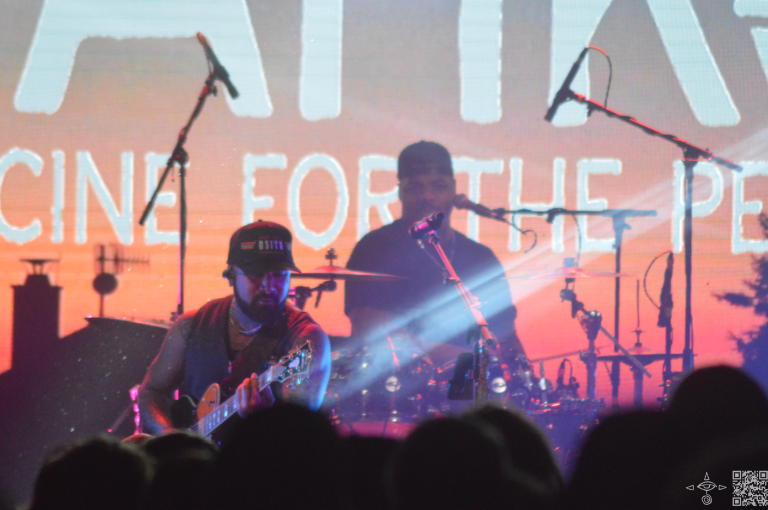 Closer up perspective of Nahko, his guitar appears to be showing a beam of light from the neck towards the upper right, A scanable code logo in the bottom right corner