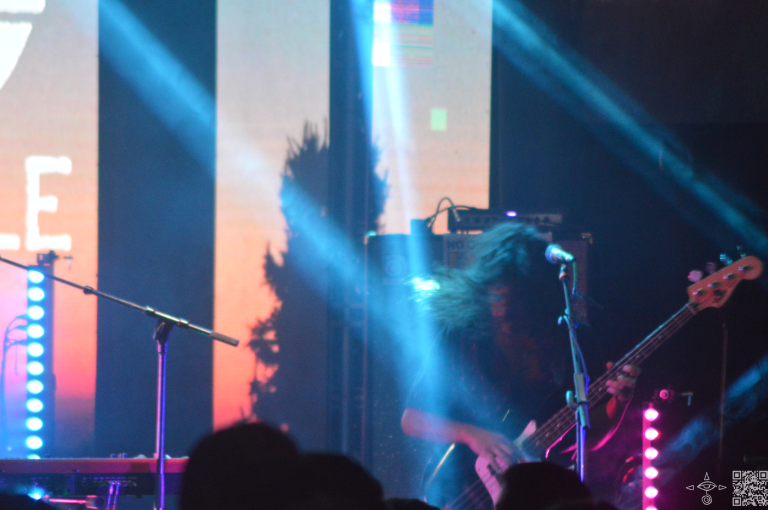 The bass player, Patricio Zuñiga Labarca, standing and playing the bass in two beam of light, long hair flying, A scanable code logo in the bottom right corner