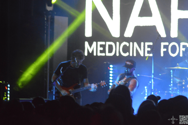 Nahko on stage Deep Blue cooling background with Nahko Logo in white letters, Lead Guitarist Joe Hall, is in the frame, A scanable code logo in the bottom right corner