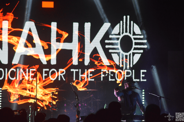 Nahko stage left at the keyboard, fire on the display behind the Nahko Logo in white font, A scanable code logo in the bottom right corner