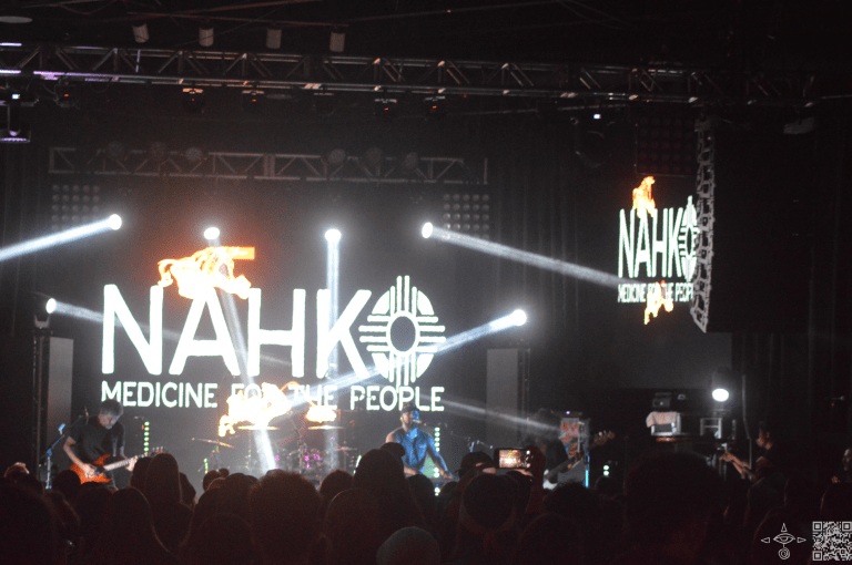 Large lights on either side of the stage, Nahko logo in white letters, with sunset colors on the stage, on the screen. A scanable code logo in the bottom right corner]