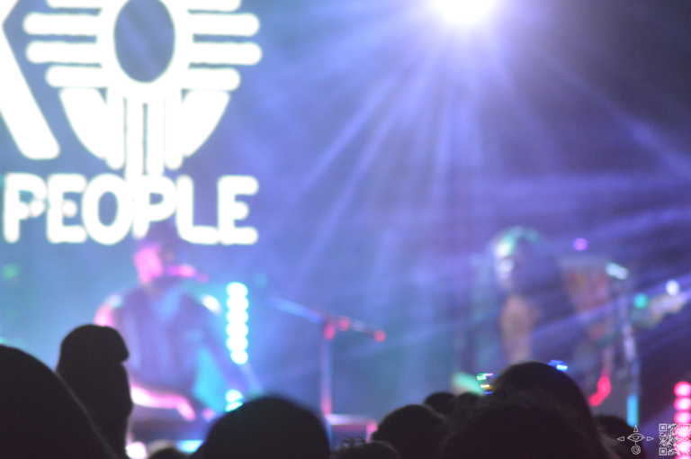 
Nahko stage left at the keyboard, fire on the display behind the Nahko Logo in white font, A blurred shot slightly zoomed out, Zia and “People” is in view from the logo above Nahko’s Head, A scanable code logo in the bottom right corner 