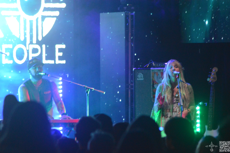 Nahko on stage, a close up perspective of Nahko at the keys stage left with Sierra Marin singing to his left. light blue and green lights like stars in the sky are projected upon the stage. A scanable code logo in the bottom right corner.