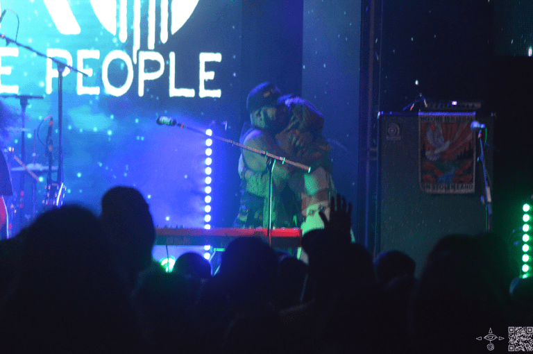 Nahko on stage, a close up perspective of Nahko  and Sierra Marin Embracing. Light blue and green lights like stars in the sky are projected upon the stage. A scanable code logo in the bottom right corner.