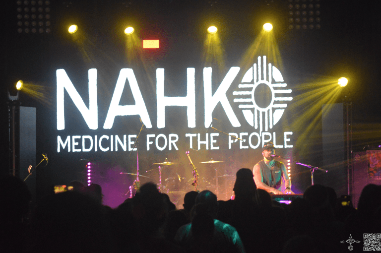Nahko Stage Left at the keys, with Graphic Display of Nahko Logo in white letters, and a silhouette of the Crowd and Beaming Yellow Light