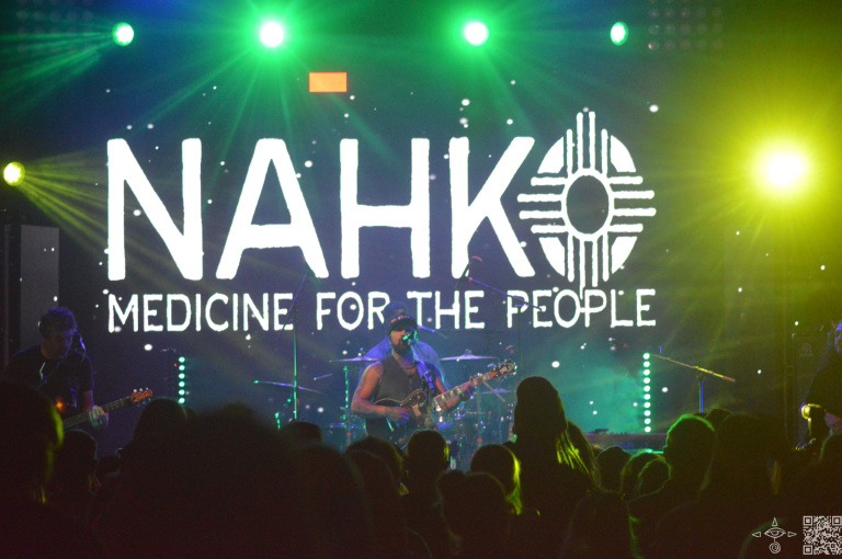 Nahko on stage, light blue, yellow and green beaming lights, Nahko logo iin white letters, the band is pictured in this photo. Medium perspective of the stage. A scanable code logo in the bottom right corner.