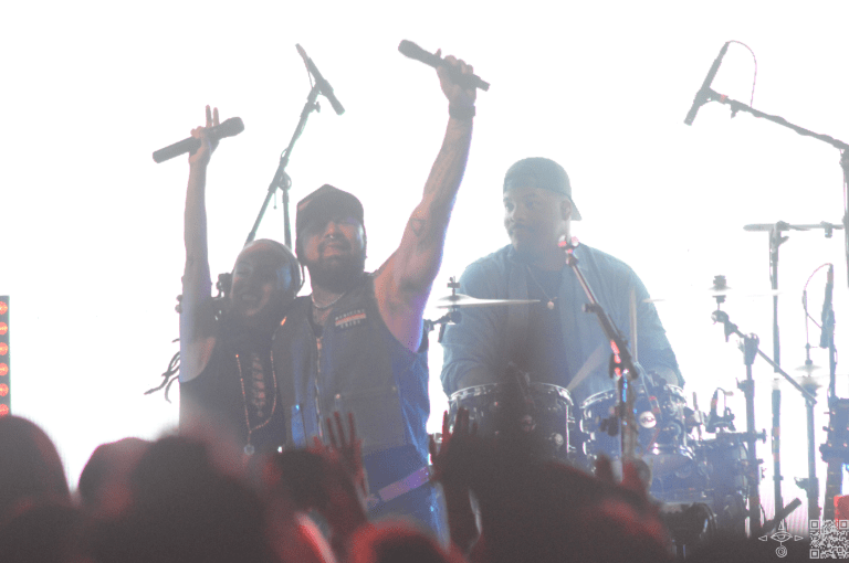 Nahko on stage with Nattali Rize, hands holding microphones up to the sky, triumphant, and the Drummer Justin Chittams, closer perspective, a light shot.