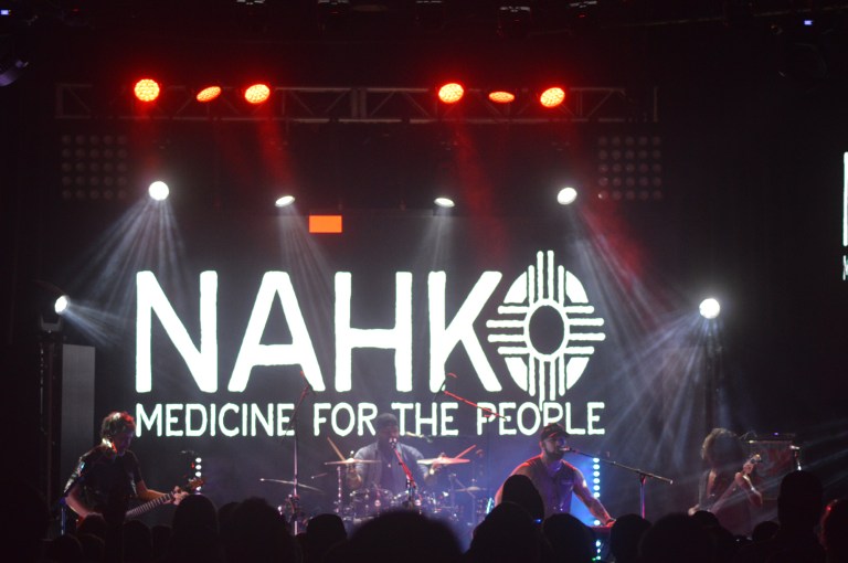 Nahko and medicine for the people logo on the display screen with Nahko written in white letters, in a Slim Angular Font with an Zia for the ”O” and Medicine for the people written below Nahko in the same font 1/6 the size of the Nahko Font, Four members of the band are on stage, A scanable code logo in the bottom right corner